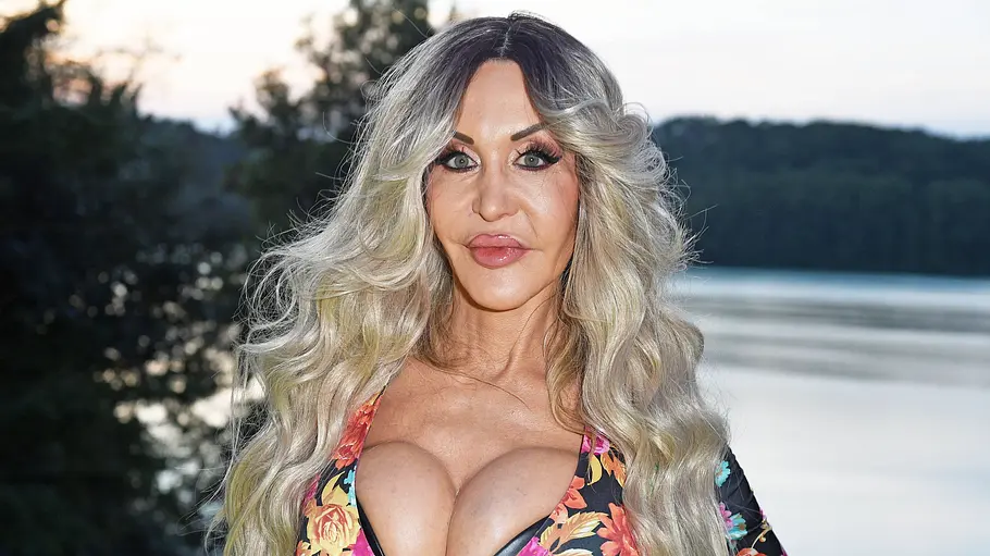 Dolly Buster steht vor einer Seelandschaft. Sie trägt ein farbenfrohes, tief ausgeschnittenes Oberteil mit Blumenmuster und blickt direkt in die Kamera. Im Hintergrund sind Bäume und Wasser zu sehen.