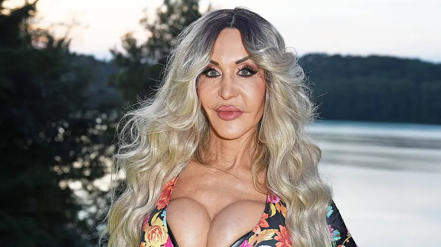 Dolly Buster im Porträt Dolly Buster posiert mit langen blonden Haaren und gemustertem Oberteil vor einem See.