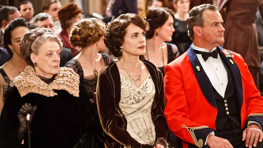 Maggie Smith, Elizabeth McGovern und Hugh Bonneville sitzen in der Serie „Downton Abbey“ in festlicher, historischer Kleidung nebeneinander.