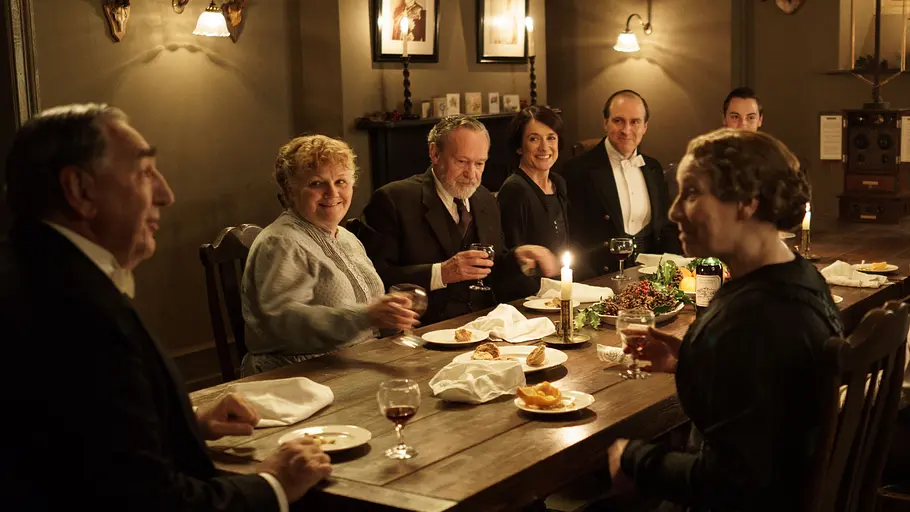 Eine Gruppe von sieben elegant gekleideten Menschen sitzt an einem festlich gedeckten Holztisch in einem stilvollen, warm beleuchteten Raum. Die Atmosphäre wirkt freundlich und gesellig, auf dem Tisch stehen Kerzen, Gläser mit Getränken und Teller mit Essen. Die Personen lachen und unterhalten sich in gemütlicher Runde. Im Hintergrund hängen Bilder und Jagdtrophäen an der Wand.