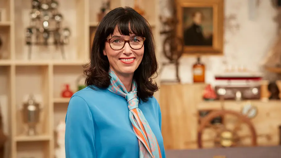 Dr. Heide Rezepa-Zabel Dr. Heide Rezepa-Zabel trägt ein blaues Kleid und ein blau-orangenes Tuch um den Hals. Sie trägt roten Lippenstift und lächelt in die Kamera.