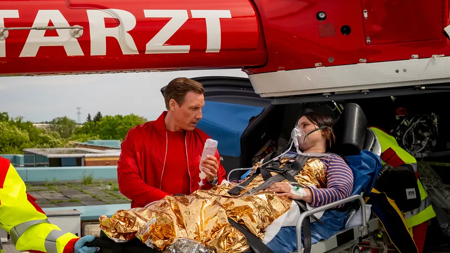 Dr. Nice: „Sommerliebe“ Die verletzte Mechanikerin Heike Kärkering (Anna Fischer) liegt auf einer Trage bedeckt mit einer Wärmedecke und wird abtransportiert. Dr. Nice (Patrick Kalupa) steht zur Seite und hält die Infusionsflasche.