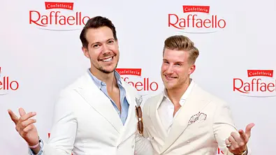 Dr. Rick und Dr. Nick in hellen Anzügen posieren mit ausgebreiteten Armen vor einer Wand mit roten Raffaello-Logos. - Foto: Imago/Panama Pictures Dr. Rick und Dr. Nick in hellen Anzügen posieren mit ausgebreiteten Armen vor einer Wand mit roten Raffaello-Logos. - Foto: Imago/Panama Pictures