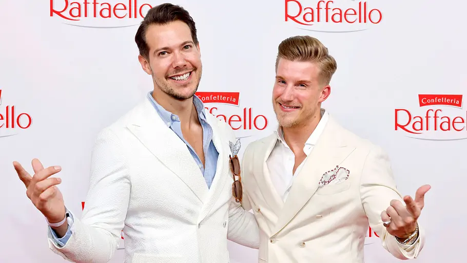 Dr. Rick und Dr. Nick Arzt Gefängnis. Dr. Rick und Dr. Nick in hellen Anzügen posieren mit ausgebreiteten Armen vor einer Wand mit roten Raffaello-Logos.