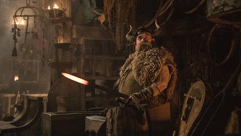 Nick Frost steht als der blonde, sowie bärtige Wikinger Grobian in grünlicher Wikinger-Montur vor einem Amboss und hat ein heißes Stück Metall in der Hand. - Foto:  IMAGO / Cinema Publishers Collection