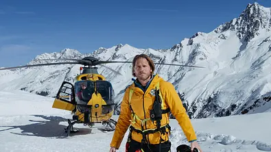 Markus Kofler steht in gelber Rettungskleidung mit Helm in der Hand vor einem Hubschrauber in verschneiter Berglandschaft. - Foto: ZDF/ Sabine Finger Fotografie Markus Kofler steht in gelber Rettungskleidung mit Helm in der Hand vor einem Hubschrauber in verschneiter Berglandschaft. - Foto: ZDF/ Sabine Finger Fotografie