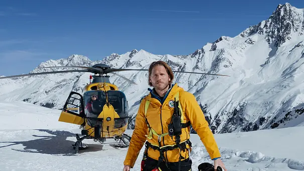 Markus Kofler steht in gelber Rettungskleidung mit Helm in der Hand vor einem Hubschrauber in verschneiter Berglandschaft. - Foto: ZDF/ Sabine Finger Fotografie Markus Kofler steht in gelber Rettungskleidung mit Helm in der Hand vor einem Hubschrauber in verschneiter Berglandschaft. - Foto: ZDF/ Sabine Finger Fotografie