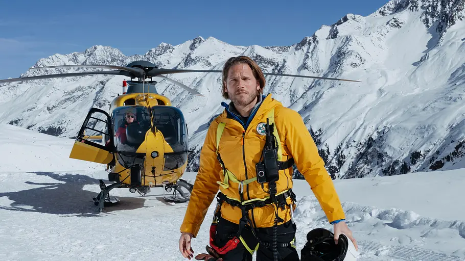 Markus Kofler Markus Kofler steht in gelber Rettungskleidung mit Helm in der Hand vor einem Hubschrauber in verschneiter Berglandschaft.