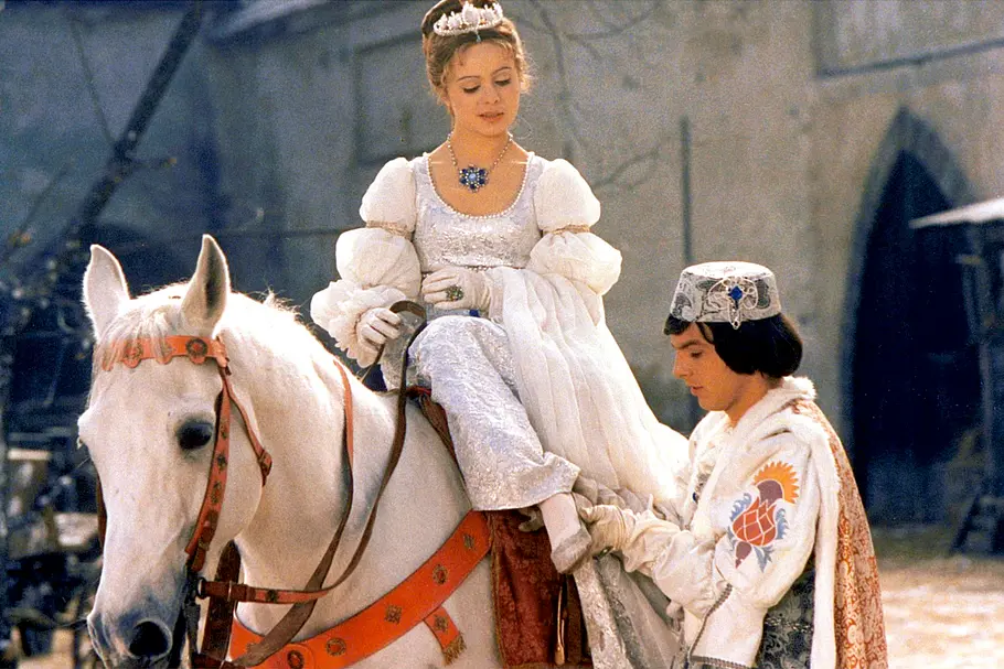 Eine junge Frau in einem prunkvollen weißen Märchenkleid mit Tiara sitzt auf einem weißen Pferd, während ein junger Mann in historischer Tracht ihr beim Aufsteigen hilft, vor der Kulisse eines Schlosses.