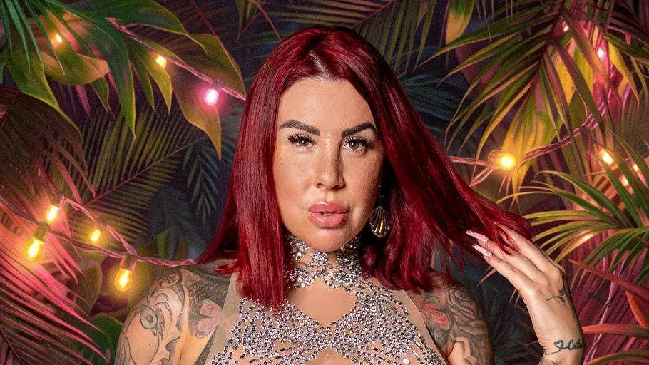 Reality-TV-Star Kate Merlan posiert für ein Promobild von „Dschungel Divas“ in einem glitzernden, transparenten Kleid mit weißem Tüllrock vor einer tropischen Kulisse mit Lichterketten und großen Palmenblättern. Sie hält eine Haarsträhne in der Hand und blickt selbstbewusst in die Kamera.