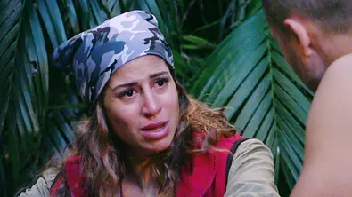 Eva mit Tarnmuster-Kopftuch und roter Weste sitzt im Dschungelcamp, schaut ernst und spricht mit einem männlichen Mitbewohner. Im Hintergrund sind grüne Blätter zu sehen. - Foto: RTL