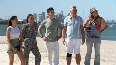 Fünf Dschungelcamp-Teilnehmer stehen am Strand vor einer Skyline und blicken gespannt nach vorn. - Foto: RTL