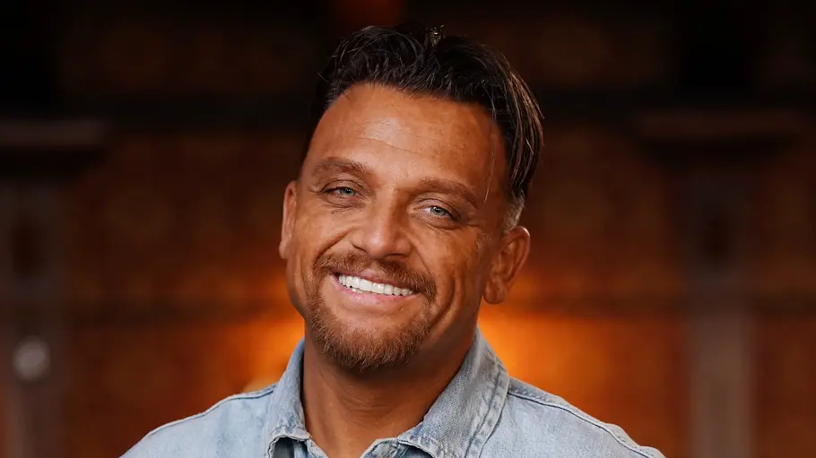 Menowin Fröhlich Menowin Fröhlich lächelt in die Kamera, er trägt ein helles Jeanshemd und steht entspannt mit den Händen in den Hosentaschen vor einem warm beleuchteten Hintergrund