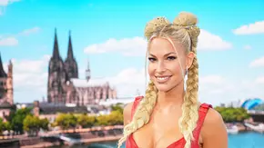 Isi Glück lächelt in einem roten, geschnürten Oberteil mit blonden Zöpfen. Im Hintergrund sind ein Fluss und eine Kathedrale zu sehen. - Foto: RTL / Stefan Gregorowius Isi Glück lächelt in einem roten, geschnürten Oberteil mit blonden Zöpfen. Im Hintergrund sind ein Fluss und eine Kathedrale zu sehen. - Foto: RTL / Stefan Gregorowius