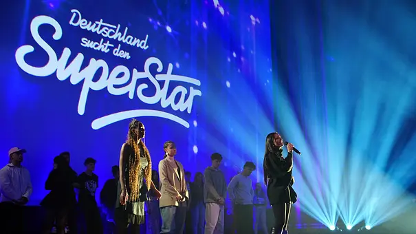 Mehrere Kandidaten stehen auf einer Bühne vor dem Schriftzug „Deutschland sucht den Superstar“, während eine Frau vorne singt. - Foto: RTL / Stefan Gregorowius Mehrere Kandidaten stehen auf einer Bühne vor dem Schriftzug „Deutschland sucht den Superstar“, während eine Frau vorne singt. - Foto: RTL / Stefan Gregorowius