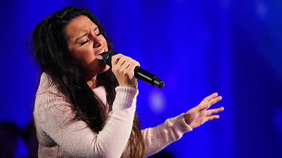 Valentina Wagner Valentina mit langen dunklen Haaren singt emotional ins Mikrofon, eine Hand ausdrucksstark erhoben, blauer Hintergrund.