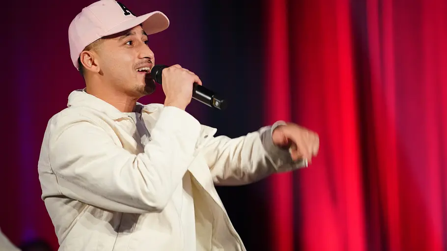 Abii Faizan Abii in heller Jacke und Kappe rappt oder singt ins Mikrofon vor rotem Bühnenvorhang.