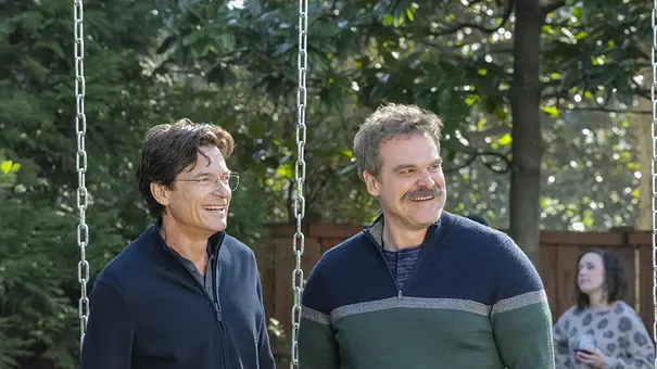 Zwei Männer stehen lächelnd nebeneinander in einem Garten neben einer Schaukel, während im Hintergrund eine Frau auf ihr Handy schaut. - Foto: Sky Atlantic