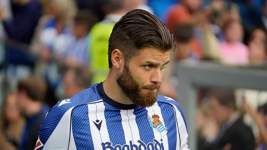 Fußballspieler von Real Sociedad im blau-weiß gestreiften Heimtrikot blickt vor einem Spiel konzentriert nach unten. Im Hintergrund sind Zuschauer und das Stadion unscharf zu erkennen.
