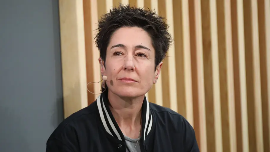 Dunja Hayali trägt eine dunkle Jacke und ein Headset-Mikrofon. Hinter ihr sind senkrechte Holzlamellen zu sehen.