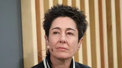 Dunja Hayali trägt eine dunkle Jacke mit gestreiftem Kragen und ein Headset-Mikrofon. Hinter ihr sind helle Holzlamellen. - Foto: IMAGO / Matthias Wehnert Dunja Hayali trägt eine dunkle Jacke mit gestreiftem Kragen und ein Headset-Mikrofon. Hinter ihr sind helle Holzlamellen. - Foto: IMAGO / Matthias Wehnert