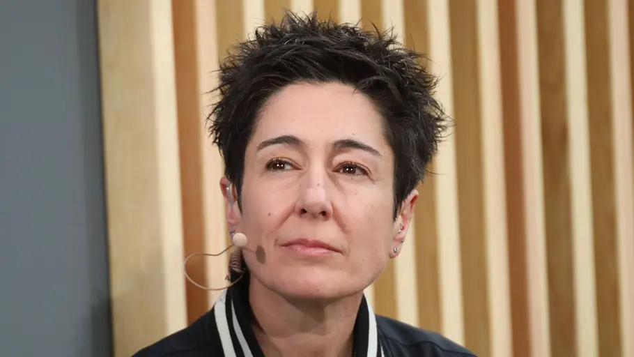 Dunja Hayali reflektiert Streit Dunja Hayali trägt eine dunkle Jacke mit gestreiftem Kragen und ein Headset-Mikrofon. Hinter ihr sind helle Holzlamellen.