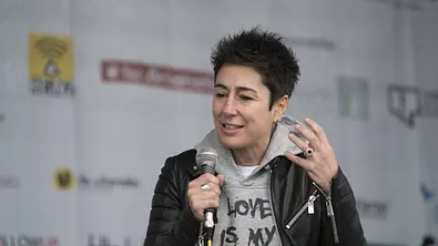 Dunja Hayali hält ein Mikrofon und gestikuliert mit erhobener Hand. Sie trägt eine schwarze Lederjacke über einem grauen Hoodie. - Foto: IMAGO / ecomedia/robert fishman