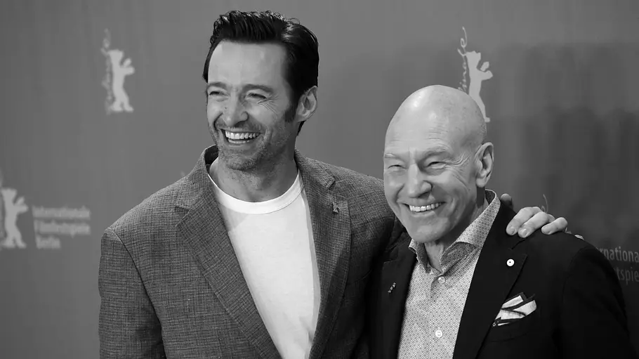Durch die „X-Men“-Filme waren Hugh Jackman und Patrick Stewart miteinander verbunden. Hugh Jackman legt lächelnd den Arm um Patrick Stewart. Beide tragen Sakkos und posieren vor einer Fotowand in Schwarz-Weiß.