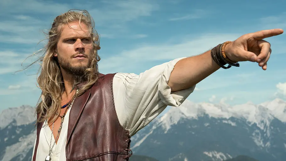 Rúrik Gíslason steht vor einer Bergkulisse und zeigt mit seinem Finger in die Ferne.