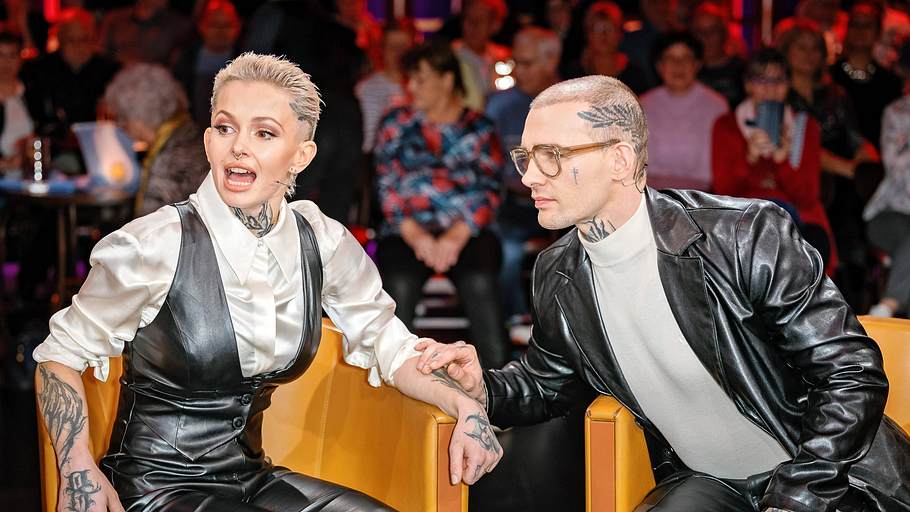Eric und Edith Stehfest sitzen auf gelben Talkshow-Stühlen. Er legt sanft die Hand auf ihren Arm. 