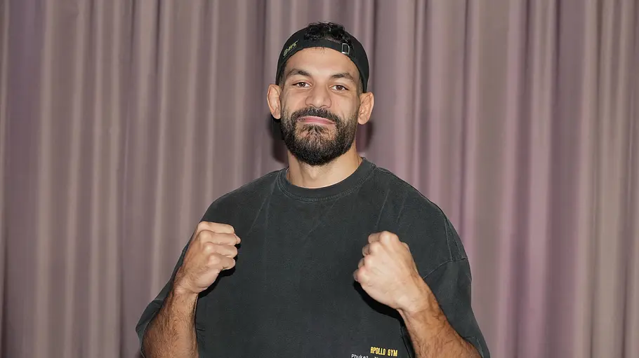 Ediz Tasci Ediz Tasci steht mit erhobenen Fäusten vor einem Vorhang. Er trägt ein dunkles T-Shirt und eine rückwärts aufgesetzte Kappe.