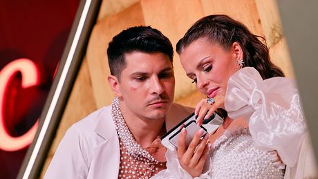 Patricija und Alexandru Ionel, beide in weißen Outfits mit Perlenbesatz, schauen am Rande von „Let’s Dance“ auf ein Handy - Foto:  IMAGO / Panama Pictures