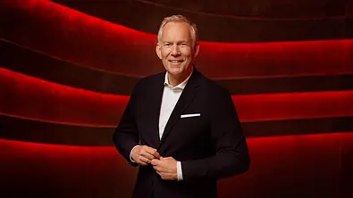 Johannes B. Kerner im dunkelblauen Anzug und weißem Hemd steht vor einem Hintergrund mit geschwungenen, leuchtend roten Linien. Er schaut in die Kamera und knöpft sein Sakko zu. - Foto: ZDF