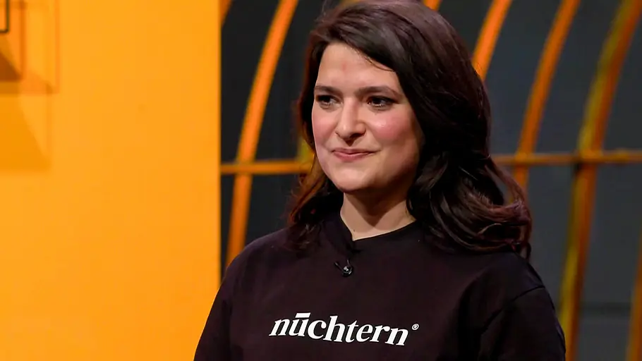 Einige Löwen stören sich am Geschmack, andere am Namen von „nüchtern“. Isabella Steiner trägt ein schwarzes Oberteil mit dem weißen Schriftzug „nüchtern“ vor einem orange-schwarzen Hintergrund.