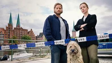 Sebastian Ströbel (l.) und Friederike Linke stehen mit einem Hund vor einem Absperrungsband der Polizei. Im Hintergrund ist das Stadtbild Lübecks zu sehen. - Foto: Sandra Hoever Photography