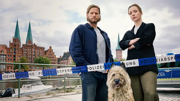 Sebastian Ströbel (l.) und Friederike Linke stehen mit einem Hund vor einem Absperrungsband der Polizei. Im Hintergrund ist das Stadtbild Lübecks zu sehen. - Foto: Sandra Hoever Photography