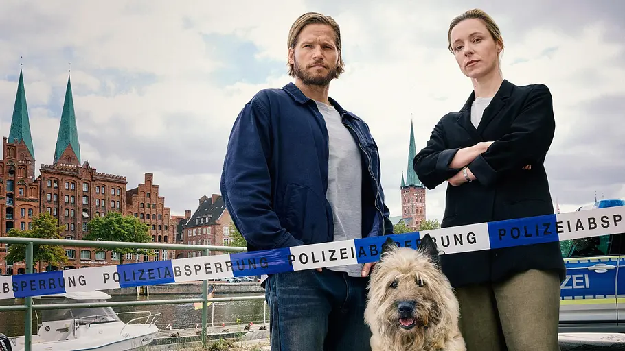 Einsatz Seeler Sebastian Ströbel (l.) und Friederike Linke stehen mit einem Hund vor einem Absperrungsband der Polizei. Im Hintergrund ist das Stadtbild Lübecks zu sehen.