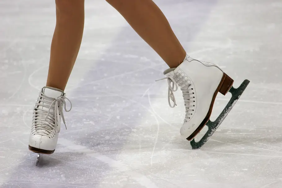Zwei Beine in weißen Eiskunstlaufschuhen sind auf einer zerkratzten Eisfläche zu sehen. Ein Schuh ist seitlich angewinkelt.