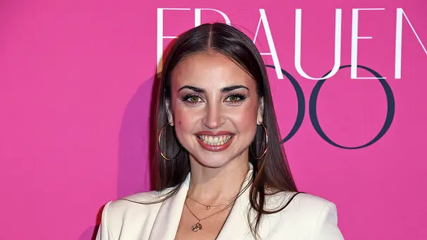 Ekaterina Leonova lächelt vor pinkem Hintergrund bei einem Event und trägt einen weißen Blazer sowie große Creolen - Foto:  IMAGO / Gartner
