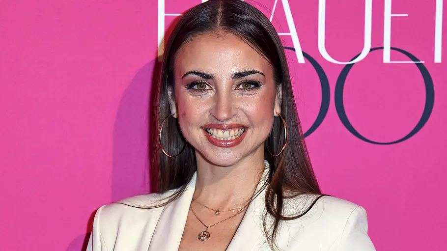 Ekaterina Leonova strahlt auf Event Ekaterina Leonova lächelt vor pinkem Hintergrund bei einem Event und trägt einen weißen Blazer sowie große Creolen