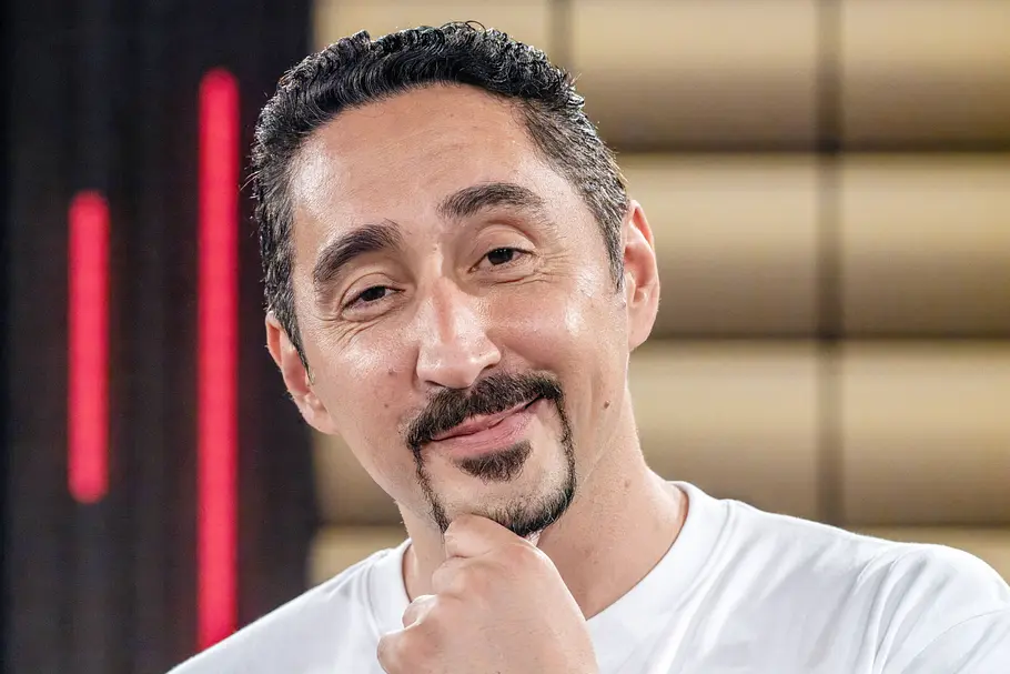 Eko Fresh Rapper Eko Fresh lächelt mit nachdenklicher Pose und auf das Kinn gestützter Hand in die Kamera. Im Hintergrund sind unscharfe Lichtelemente eines TV-Studios zu sehen.