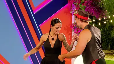 Elena und ein Kandidat tanzen Hand in Hand vor einer bunt beleuchteten Kulisse mit Blumen und Lichterketten. - Foto: RTL