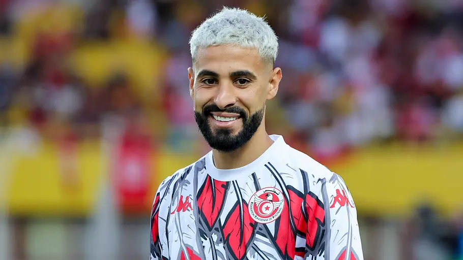 Ein lächelnder Fußballspieler der tunesischen Nationalmannschaft trägt ein auffällig gemustertes Aufwärmtrikot und blickt vor dem Spiel in die Kamera. Im Hintergrund ist das Stadion unscharf zu sehen.