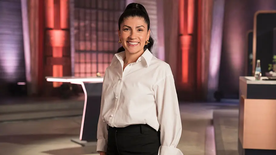 Elif Oskan Elif Oskan steht lächelnd in weißer Bluse und schwarzer Hose vor Arbeitsflächen in einem rot und violett beleuchteten Studio.