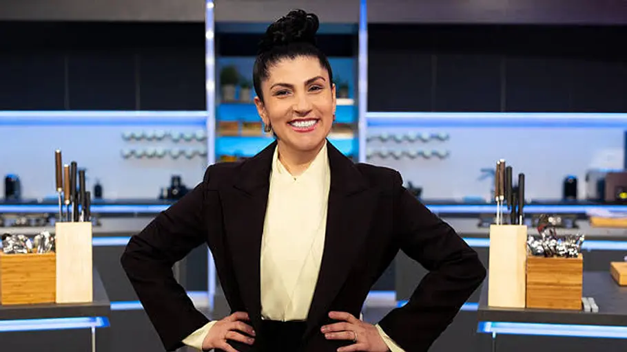 Elif Oskan The Taste Elif Oskan steht lächelnd mit den Händen in den Hüften in einer Küche. Sie trägt einen schwarzen Blazer und eine helle Bluse.