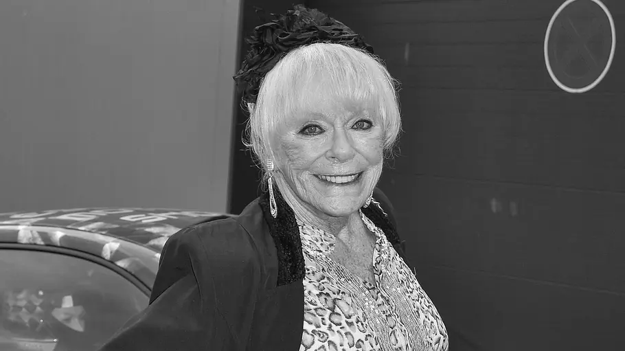 Elke Sommer gestorben Elke Sommer lächelt mit einer Hand an der Hüfte vor einem Rennwagen. Sie trägt eine gemusterte Bluse und eine dunkle Jacke.