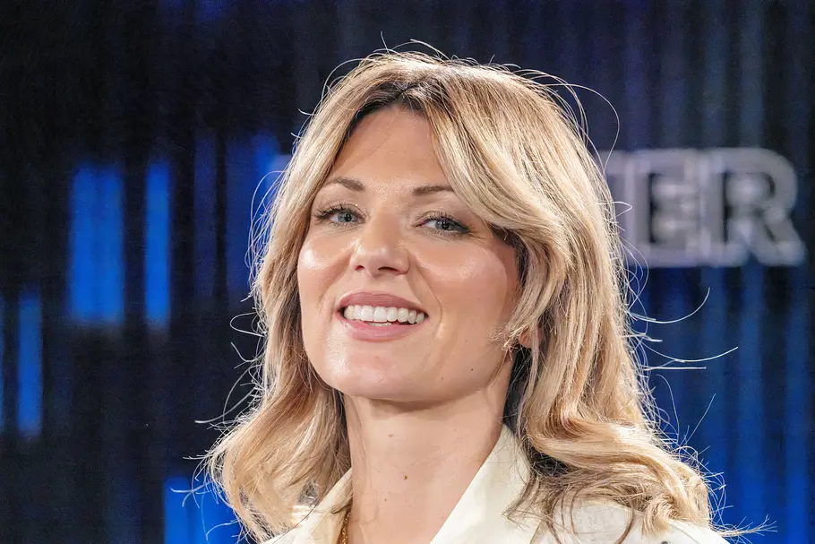 Ella Endlich Ella Endlich lächelt in einem hellen Blazer in die Kamera. Die Sängerin trägt ihr schulterlanges blondes Haar offen, im Hintergrund ist ein unscharfes TV-Studio mit dunklen Vorhängen und blauen Lichtelementen zu sehen.