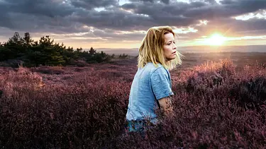 Elodie sitzt im hellblauen Oberteil in blühender Heide und blickt zur Seite. Hinter ihr steht die Sonne tief unter dunklen Wolken. - Foto: © NDR/Frizzi Kurkhaus/Viktoria Grunjajew Elodie sitzt im hellblauen Oberteil in blühender Heide und blickt zur Seite. Hinter ihr steht die Sonne tief unter dunklen Wolken. - Foto: © NDR/Frizzi Kurkhaus/Viktoria Grunjajew