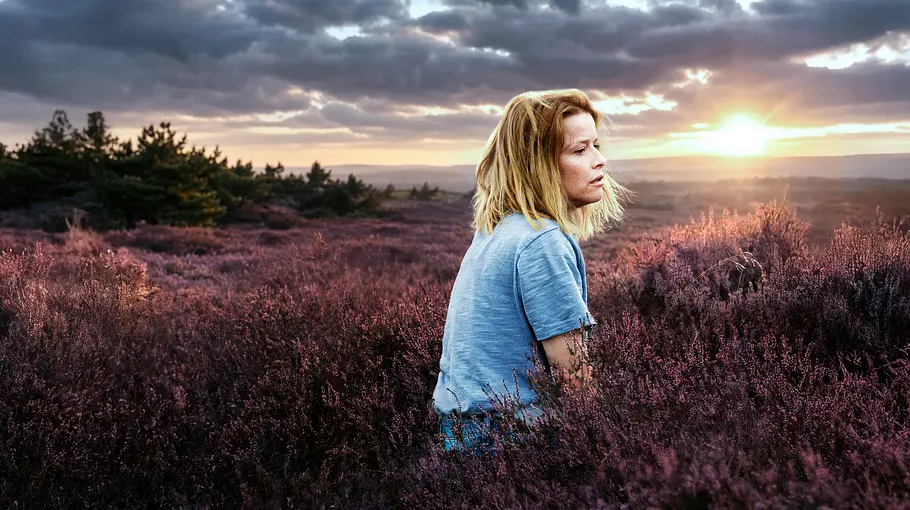 Elodie sitzt im hellblauen Oberteil in blühender Heide und blickt zur Seite. Hinter ihr steht die Sonne tief unter dunklen Wolken.