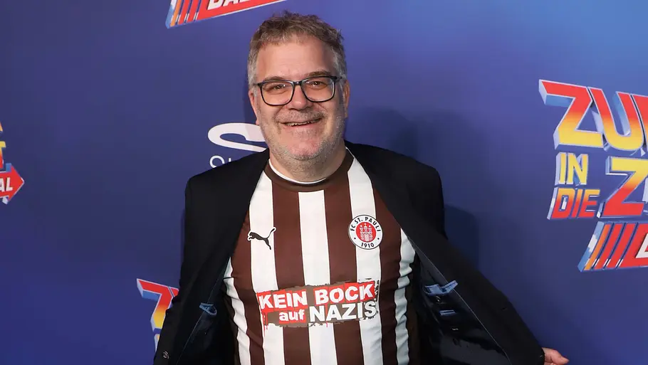 Elton Elton mit Brille, Jeans und einem braun-weiß gestreiften Fußballtrikot mit der Aufschrift „Kein Bock auf Nazis“ öffnet seine Jacke und steht vor einer blauen Fotowand.